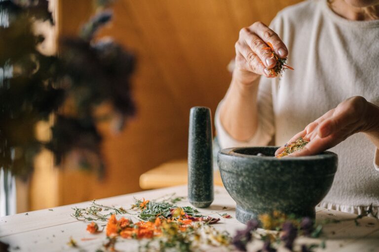 Close-up of hands grinding dried herbs and flowers in a mortar and pestle on a wooden table.